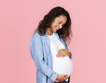 Uśmiechnięta kobieta w ciąży stojącą na tle jasnoróżowej ściany. Ma na sobie białą koszulkę i jasnoniebieską koszulę, jedną ręką podtrzymuje brzuch, a drugą delikatnie go dotyka, patrząc na niego z czułością.