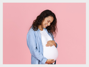 Uśmiechnięta kobieta w ciąży stojącą na tle jasnoróżowej ściany. Ma na sobie białą koszulkę i jasnoniebieską koszulę, jedną ręką podtrzymuje brzuch, a drugą delikatnie go dotyka, patrząc na niego z czułością.